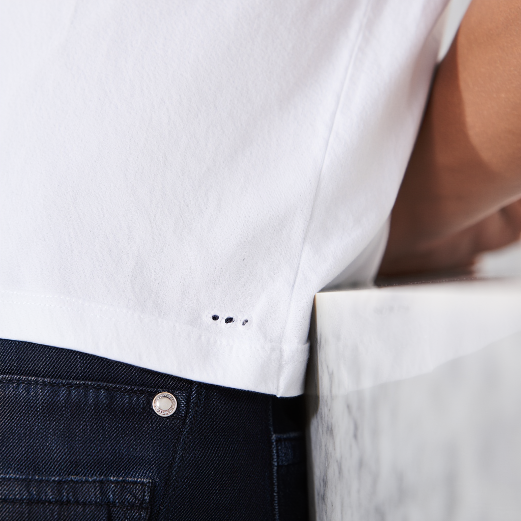 Close macro of a cotton T-shirt hem with 2–3 tiny holes forming where the fabric rubs against a metal jean button and kitchen counter edge.